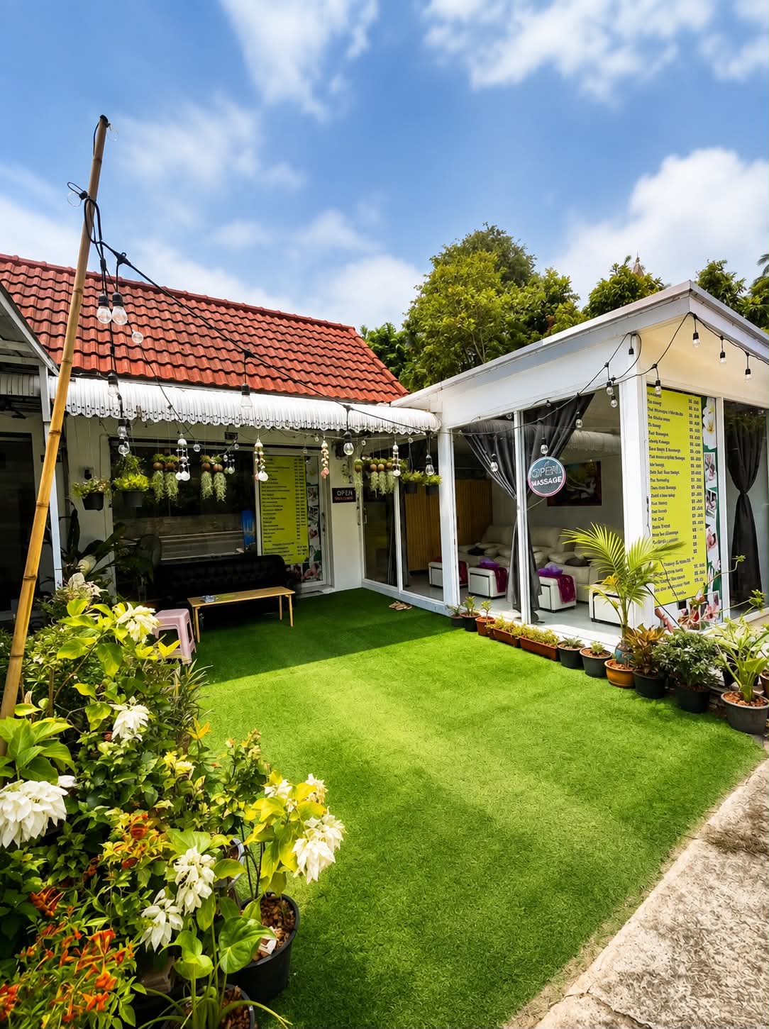 The salon by day: the green courtyard between the two buildings, the treatment boards in the windows, potted plants along the path and the massage rooms open behind the glass.