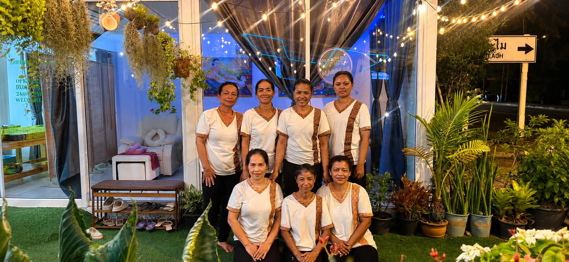 Seven of the Sabai Siam therapists together outside the salon in the evening, the neon sign behind them.